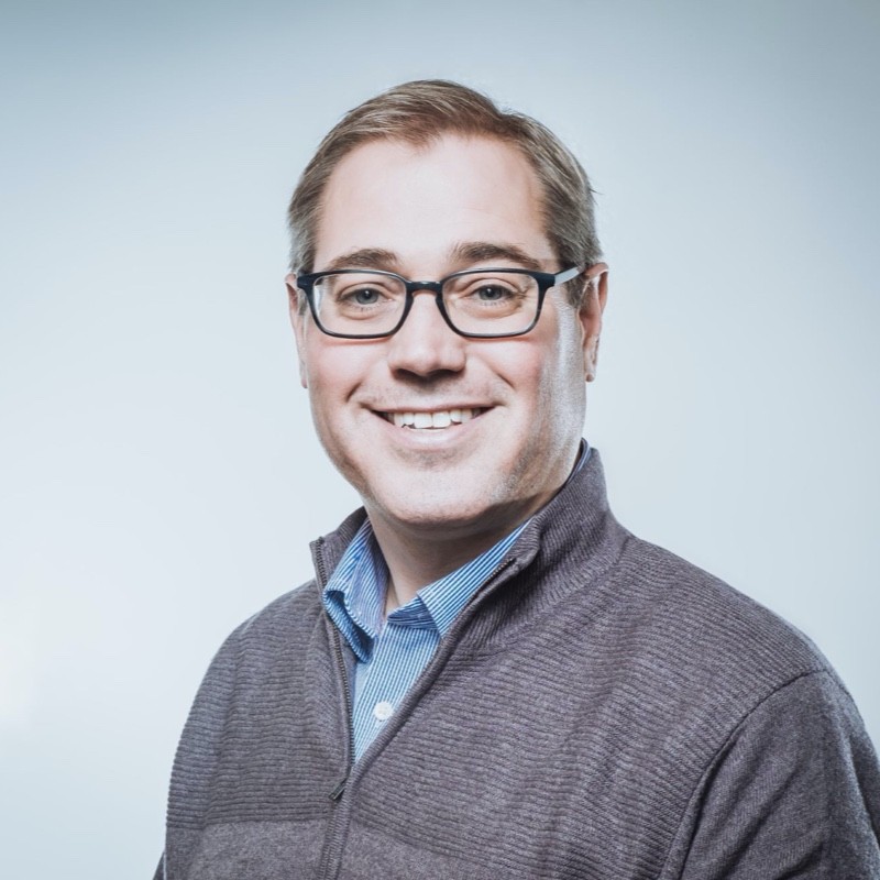 White man with glasses smiling in front of a blue background